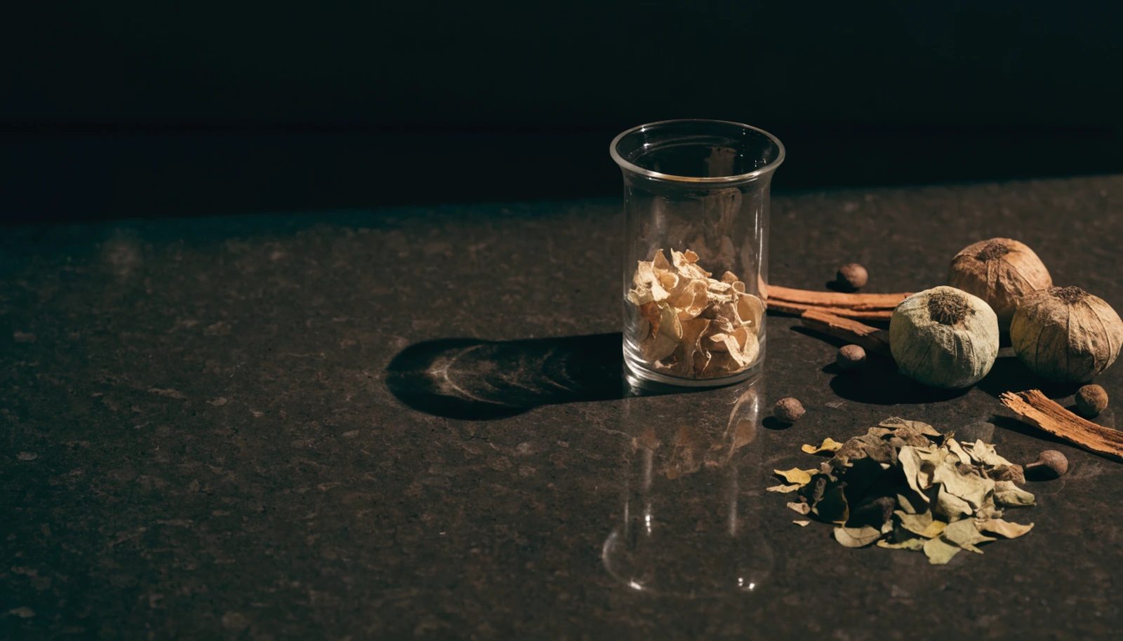 Dried botanicals and a glass beaker of petals arranged on a polished dark stone bench.