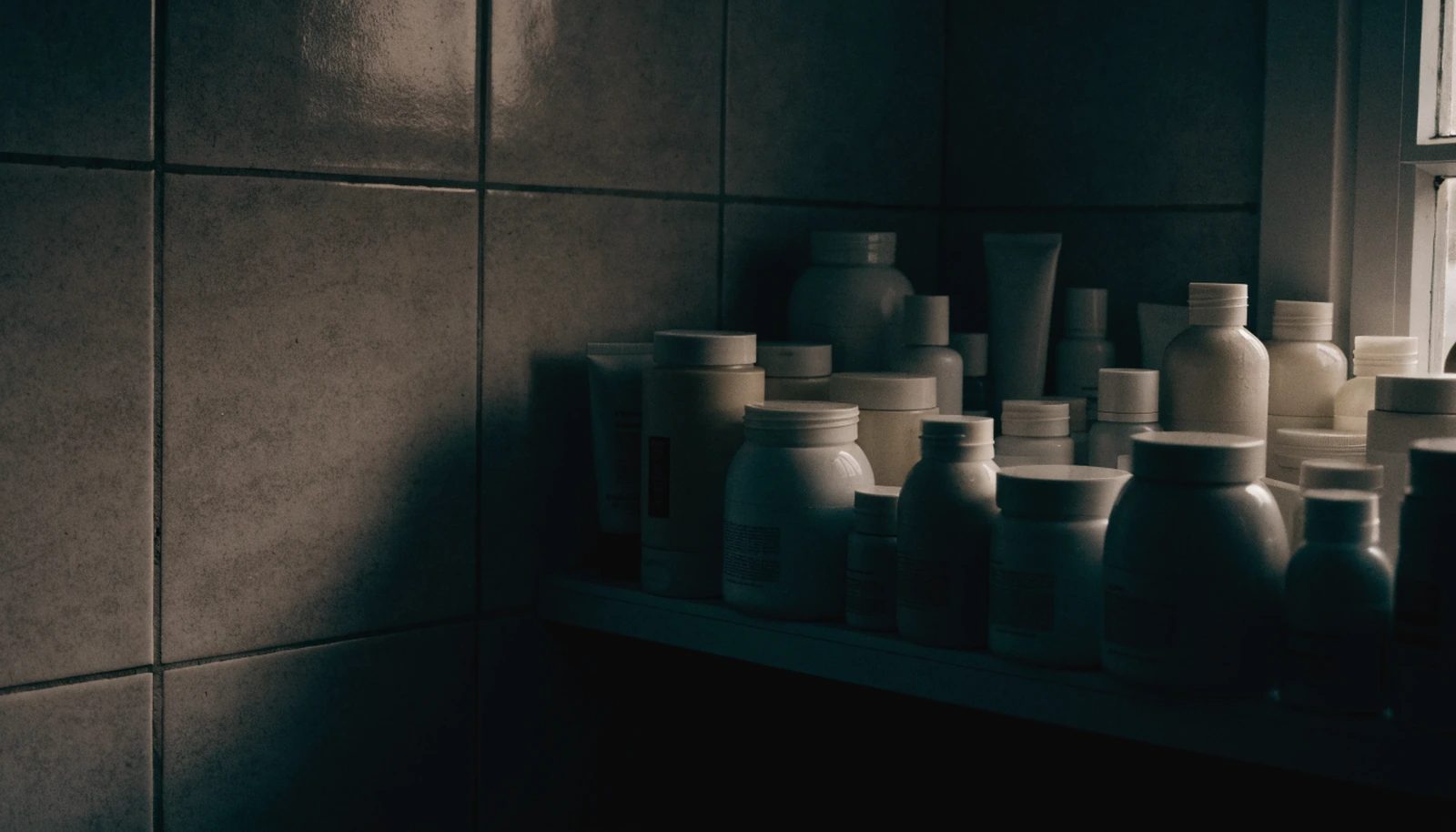 A crowded bathroom shelf of unlabelled jars and bottles in dim morning light.