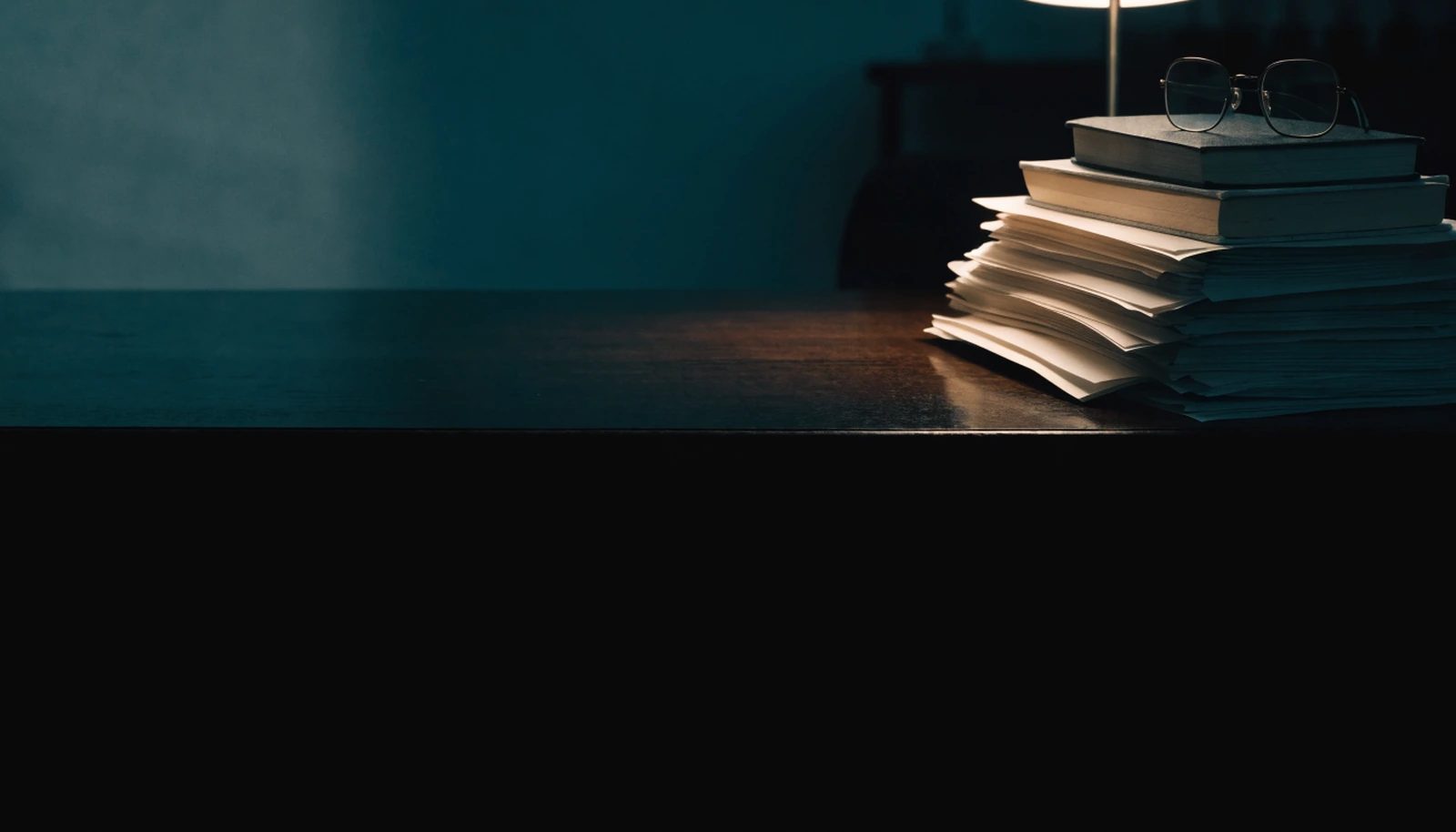 A stack of loose paper and two closed books on a dark desk beside a low lamp.